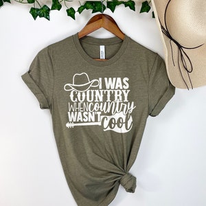 May include: A green t-shirt with the text "I was country when country wasn't cool" in white. The text is accompanied by a graphic of a cowboy hat and a guitar.