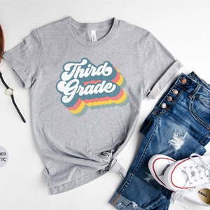 May include: A heather grey t-shirt with a retro-style rainbow graphic that says "Third Grade".