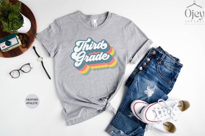 May include: A heather grey t-shirt with a retro-style rainbow graphic that says "Third Grade".