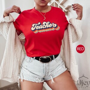 Puede incluir: Camiseta roja con un dise&ntilde;o retro de arco&iacute;ris que dice "Teacher".