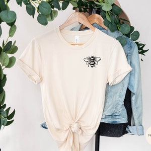 May include: A beige t-shirt with a black bee graphic on the left chest. The shirt is knotted at the bottom and hanging on a wooden hanger. The word "NATURAL" is printed in black on a beige circle in the lower right corner of the image.