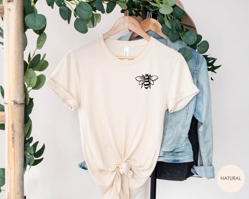 May include: A beige t-shirt with a black bee graphic on the left chest. The shirt is knotted at the bottom and hanging on a wooden hanger. The word "NATURAL" is printed in black on a beige circle in the lower right corner of the image.