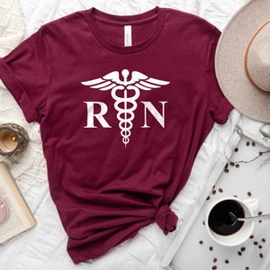 May include: A maroon t-shirt with a white medical symbol and the letters "RN" printed on the front.