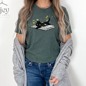 Puede incluir: Una camiseta verde oscuro con un gato negro leyendo un libro rodeado de flores. El gato est&aacute; mirando el libro. La camiseta tiene un aspecto desgastado.
