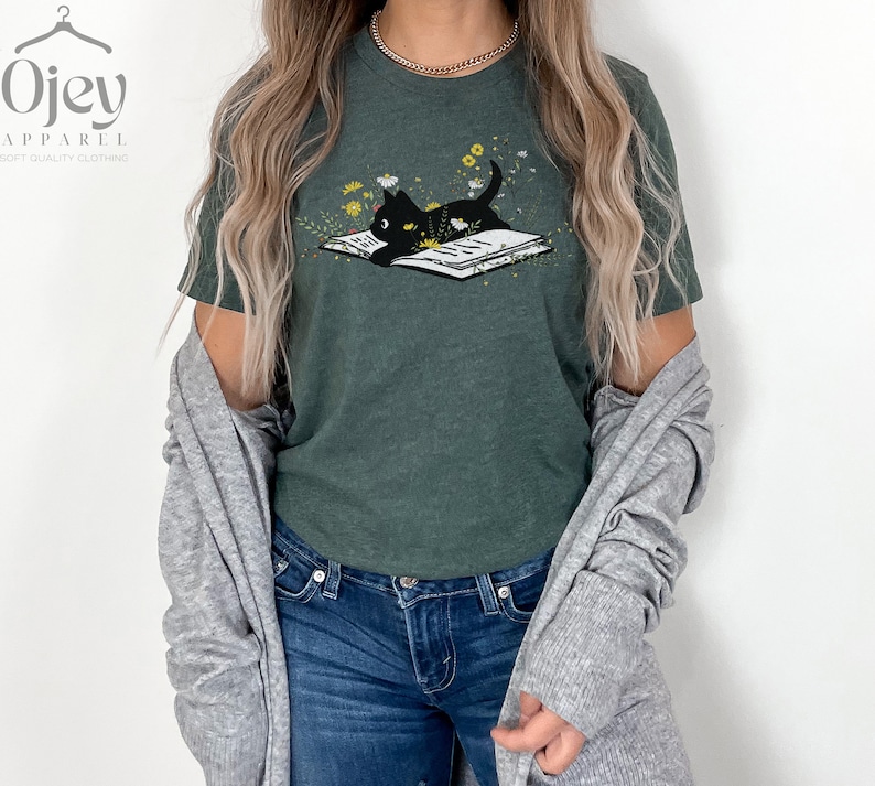 Puede incluir: Una camiseta verde oscuro con un gato negro leyendo un libro rodeado de flores. El gato est&aacute; mirando el libro. La camiseta tiene un aspecto desgastado.
