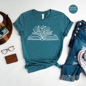 K&ouml;nnte beinhalten: Ein blaues T-Shirt mit einem wei&szlig;en Umriss eines offenen Buches, aus dem Blumen wachsen. Das Shirt liegt flach auf einer wei&szlig;en Oberfl&auml;che mit einer Brille, einer braunen Geldb&ouml;rse, einer Kamera und einer Topfpflanze. Eine blaue Jeans und wei&szlig;e Turnschuhe sind ebenfalls auf dem Bild zu sehen. Der Text "HEATHER DEEP TEAL" befindet sich in einem Kreis oben rechts im Bild.