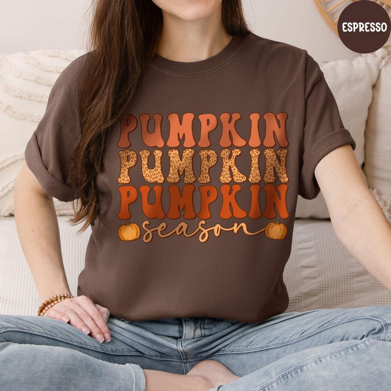 May include: A brown t-shirt with the words "PUMPKIN PUMPKIN PUMPKIN" in orange and leopard print, with "season" in cursive below. Two small orange pumpkins flank the word "season". The shirt is a solid brown color.