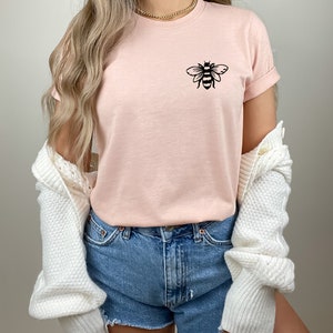 May include: A pink t-shirt with a black bee graphic on the front. The shirt is being worn with denim shorts and a white cardigan.