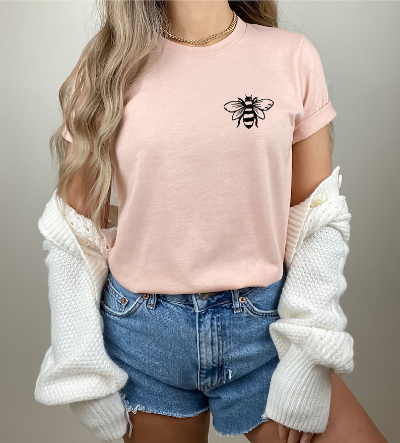 May include: A pink t-shirt with a black bee graphic on the front. The shirt is being worn with denim shorts and a white cardigan.