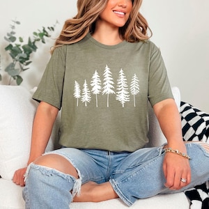 May include: A woman wearing a green t-shirt with a white graphic of a forest on it. She is sitting on a white couch with a black and white blanket beside her.