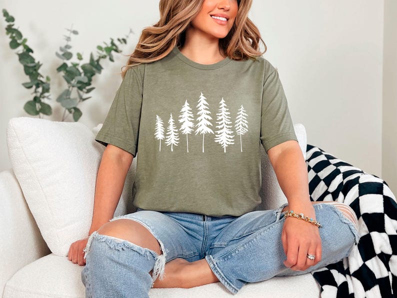 May include: A woman wearing a green t-shirt with a white graphic of a forest on it. She is sitting on a white couch with a black and white blanket beside her.