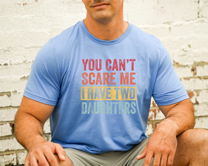 Puede incluir: Una camiseta azul claro con un gr&aacute;fico de estilo vintage que dice "You can't scare me I have two daughters".