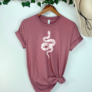 May include: A mauve t-shirt with a white snake design featuring stars and moons. The snake is curled up in a circle.