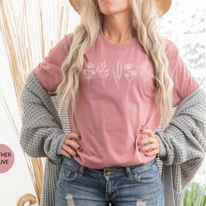 May include: A heather mauve t-shirt with a white floral design. The shirt is being worn by a person with long blonde hair and a straw hat. The text "Ojev Apparel" is visible in the background.