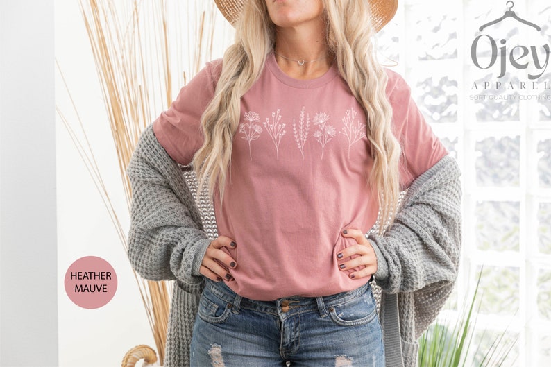 May include: A heather mauve t-shirt with a white floral design. The shirt is being worn by a person with long blonde hair and a straw hat. The text "Ojev Apparel" is visible in the background.