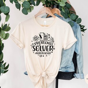 May include: A beige t-shirt with a black graphic that says "Problem Solver" with math symbols and the hashtag "#mathteacher".