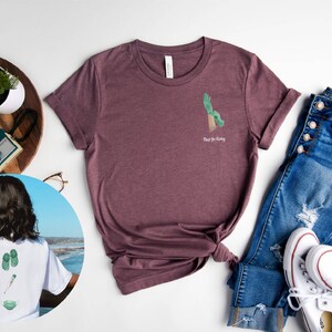 May include: A maroon t-shirt with a green hand graphic and the text "Rise & Grind" on the front. The shirt is laid out on a white surface with a pair of blue jeans, white sneakers, and a potted plant.