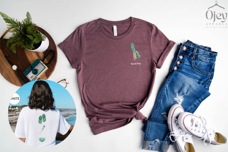 May include: A maroon t-shirt with a green hand graphic and the text "Rise & Grind" on the front. The shirt is laid out on a white surface with a pair of blue jeans, white sneakers, and a potted plant.