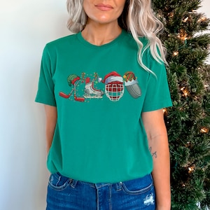 May include: A green t-shirt with a Christmas-themed hockey design. The design features a hockey stick, skates, a hockey helmet, and a Santa hat.