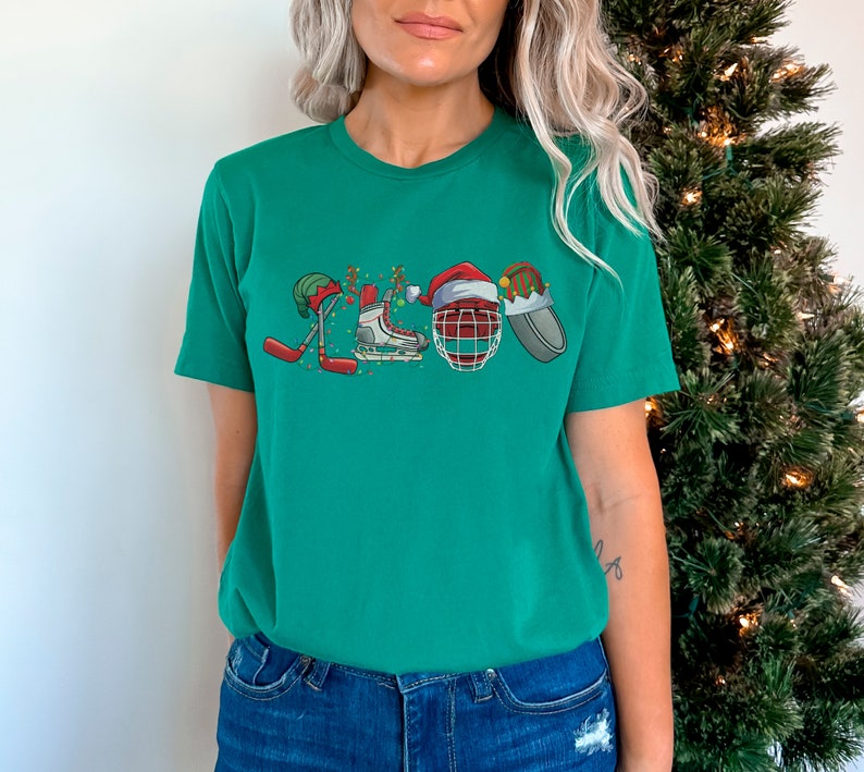 May include: A green t-shirt with a Christmas-themed hockey design. The design features a hockey stick, skates, a hockey helmet, and a Santa hat.
