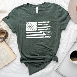 K&ouml;nnte beinhalten: Ein T-Shirt in der Farbe Heidewaldgr&uuml;n mit einem wei&szlig;en, abgenutzten Design einer amerikanischen Flagge. Die Flagge besteht aus fischereilichen Gegenst&auml;nden, darunter eine Angelrute, ein K&ouml;der, ein Fisch und ein Boot.