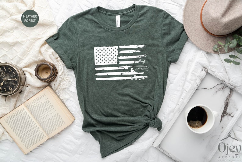 K&ouml;nnte beinhalten: Ein T-Shirt in der Farbe Heidewaldgr&uuml;n mit einem wei&szlig;en, abgenutzten Design einer amerikanischen Flagge. Die Flagge besteht aus fischereilichen Gegenst&auml;nden, darunter eine Angelrute, ein K&ouml;der, ein Fisch und ein Boot.