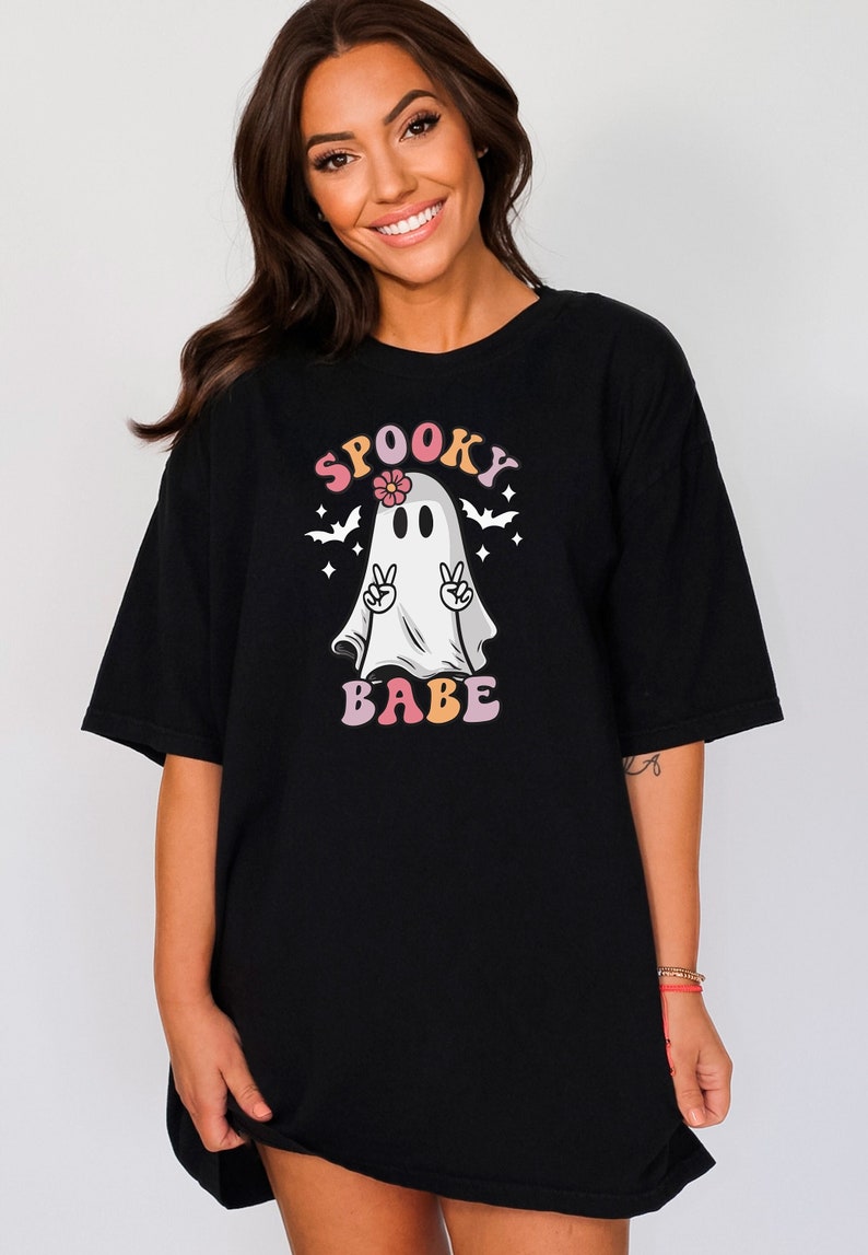 May include: Black oversized t-shirt with a graphic of a ghost wearing a flower and making a peace sign. The text "SPOOKY BABE" is written in rainbow letters.