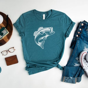May include: A teal heather deep teal t-shirt featuring a white silhouette of a bass fish leaping out of the water. The shirt is laid flat on a white surface with a pair of jeans, a pair of white trainers, a camera, a watch, and a pair of glasses.