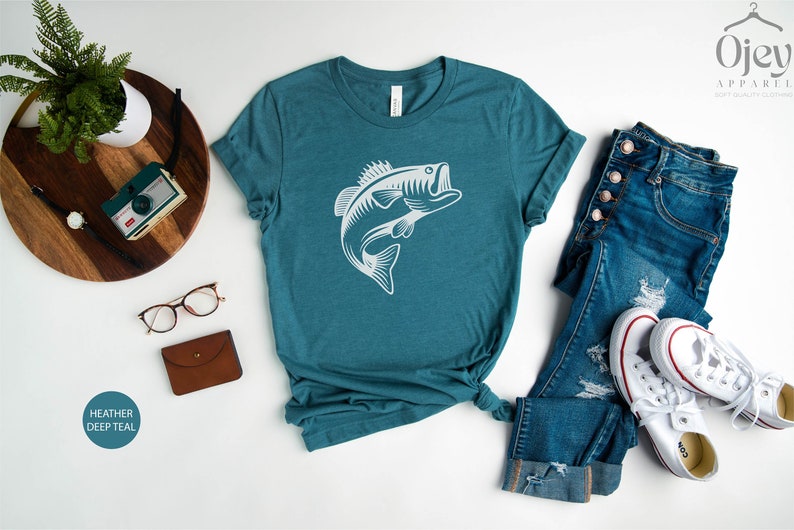May include: A teal heather deep teal t-shirt featuring a white silhouette of a bass fish leaping out of the water. The shirt is laid flat on a white surface with a pair of jeans, a pair of white trainers, a camera, a watch, and a pair of glasses.