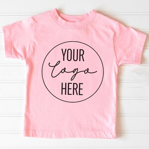 May include: A pink short-sleeved t-shirt with a black circle on the front that says "Your Logo Here" in a handwritten font.