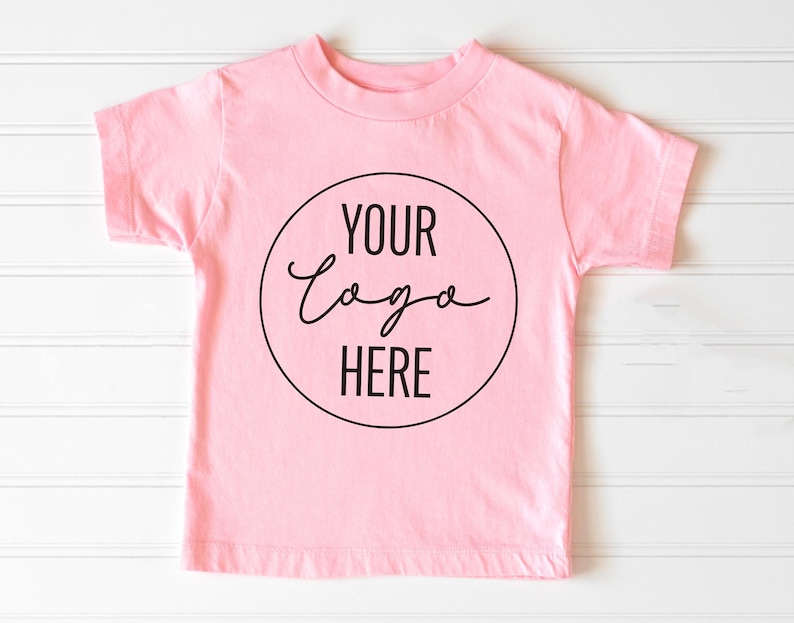 May include: A pink short-sleeved t-shirt with a black circle on the front that says "Your Logo Here" in a handwritten font.