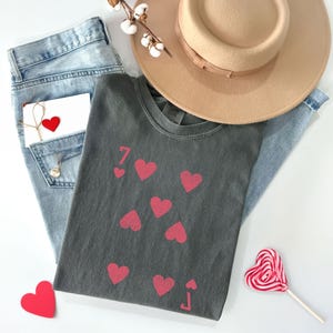 May include: A casual flat lay featuring a gray t-shirt with a playing card design of seven hearts and a jack of hearts. Light blue jeans, a tan hat, a heart-shaped lollipop, and a small card with a red heart complete the arrangement.