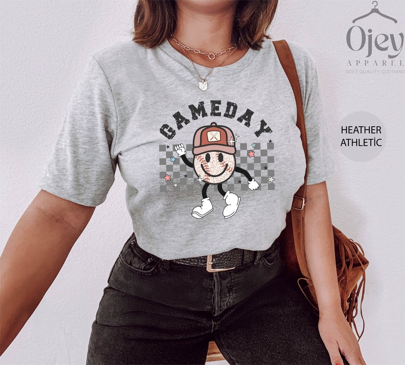 May include: A heather grey t-shirt with a graphic of a smiling baseball with a red hat and the text "GAMEDAY" in black letters.