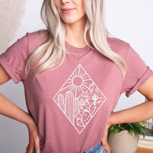 May include: A heather mauve t-shirt with a white graphic of a desert scene with cacti and a sun. The graphic is inside a diamond shape. The text "Ojev Apparel Soft Quality Clothing" and "Heather Mauve" are visible in the image.