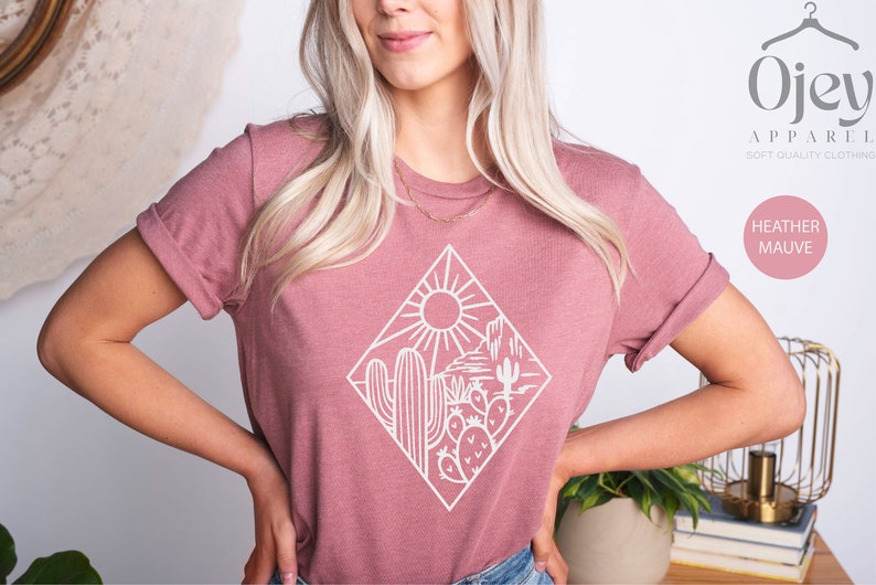 May include: A heather mauve t-shirt with a white graphic of a desert scene with cacti and a sun. The graphic is inside a diamond shape. The text "Ojev Apparel Soft Quality Clothing" and "Heather Mauve" are visible in the image.