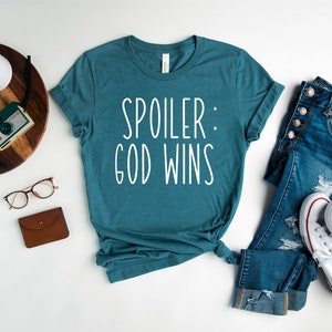 May include: Teal blue t-shirt with white text that reads "Spoiler: God Wins". The shirt is laid flat on a white surface with a pair of jeans, a pair of white trainers, a pair of glasses, a brown wallet, and a vintage camera on a wooden tray.