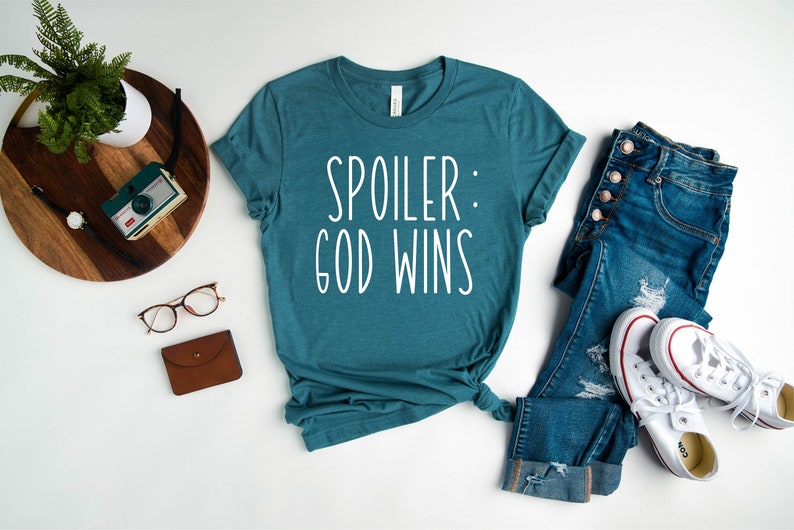 May include: Teal blue t-shirt with white text that reads "Spoiler: God Wins". The shirt is laid flat on a white surface with a pair of jeans, a pair of white trainers, a pair of glasses, a brown wallet, and a vintage camera on a wooden tray.