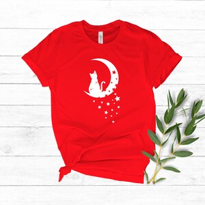 May include: Red t-shirt with a white silhouette of a cat sitting on a crescent moon with stars.