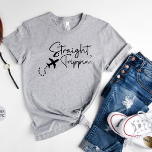 May include: A heather grey t-shirt with the text "Straight, Trippin'" and a small airplane graphic. The shirt is knotted at the waist and is paired with blue jeans and white sneakers.