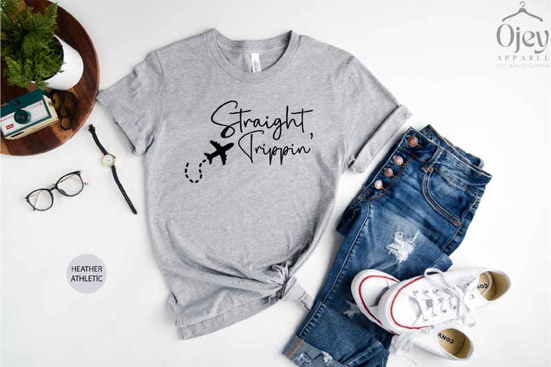 May include: A heather grey t-shirt with the text "Straight, Trippin'" and a small airplane graphic. The shirt is knotted at the waist and is paired with blue jeans and white sneakers.