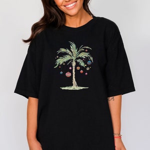 K&ouml;nnte beinhalten: Schwarzes Oversize-T-Shirt mit einem gr&uuml;nen Palmenbaum, der mit bunten Weihnachtskugeln geschm&uuml;ckt ist.