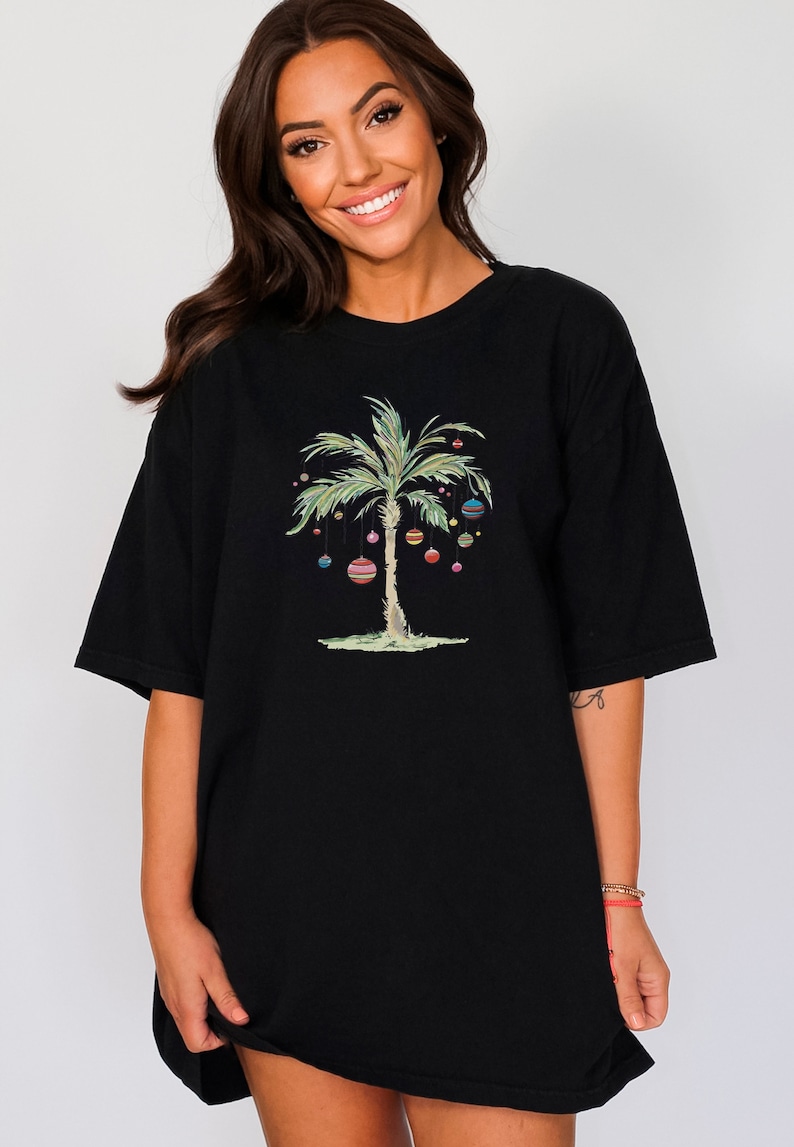 K&ouml;nnte beinhalten: Schwarzes Oversize-T-Shirt mit einem gr&uuml;nen Palmenbaum, der mit bunten Weihnachtskugeln geschm&uuml;ckt ist.
