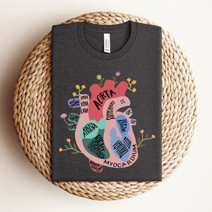May include: A dark gray t-shirt with a colorful illustration of a human heart. The heart is labeled with the names of its parts, including the aorta, right atrium, right ventricle, left atrium, left ventricle, and myocardium. The illustration is surrounded by small flowers.
