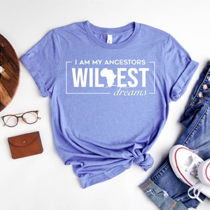 Peut inclure: Un t-shirt bleu heather columbia avec le texte "I am my ancestors wildest dreams" en blanc avec un contour du continent africain. Le t-shirt est nou&eacute; et dispos&eacute; sur une surface blanche avec une paire de lunettes, un portefeuille marron, un appareil photo et une paire de jeans et de baskets blanches.