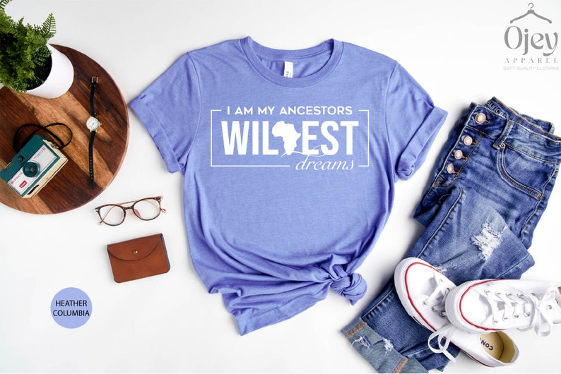 Peut inclure: Un t-shirt bleu heather columbia avec le texte "I am my ancestors wildest dreams" en blanc avec un contour du continent africain. Le t-shirt est nou&eacute; et dispos&eacute; sur une surface blanche avec une paire de lunettes, un portefeuille marron, un appareil photo et une paire de jeans et de baskets blanches.