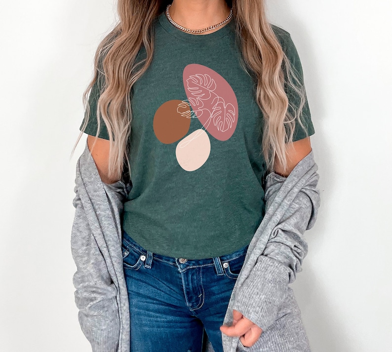 Puede incluir: Una camiseta verde oscuro con un dise&ntilde;o gr&aacute;fico de formas abstractas en rosa, marr&oacute;n y blanco. El dise&ntilde;o presenta una hoja estilizada.