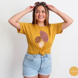 Puede incluir: Una camiseta amarilla mostaza con un gr&aacute;fico de un dise&ntilde;o abstracto marr&oacute;n, rosa y blanco con un dibujo lineal de una hoja. El texto "MUSTARD" es visible en la esquina inferior derecha de la imagen.