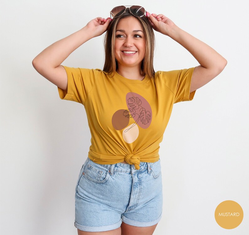Puede incluir: Una camiseta amarilla mostaza con un gr&aacute;fico de un dise&ntilde;o abstracto marr&oacute;n, rosa y blanco con un dibujo lineal de una hoja. El texto "MUSTARD" es visible en la esquina inferior derecha de la imagen.