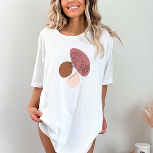 Puede incluir: Camiseta blanca de gran tama&ntilde;o con un dise&ntilde;o gr&aacute;fico de formas abstractas en rosa, marr&oacute;n y beige con un dibujo lineal de una hoja.