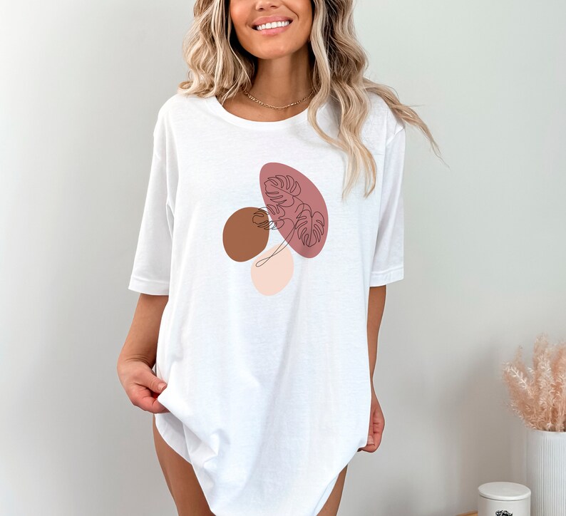 Puede incluir: Camiseta blanca de gran tama&ntilde;o con un dise&ntilde;o gr&aacute;fico de formas abstractas en rosa, marr&oacute;n y beige con un dibujo lineal de una hoja.
